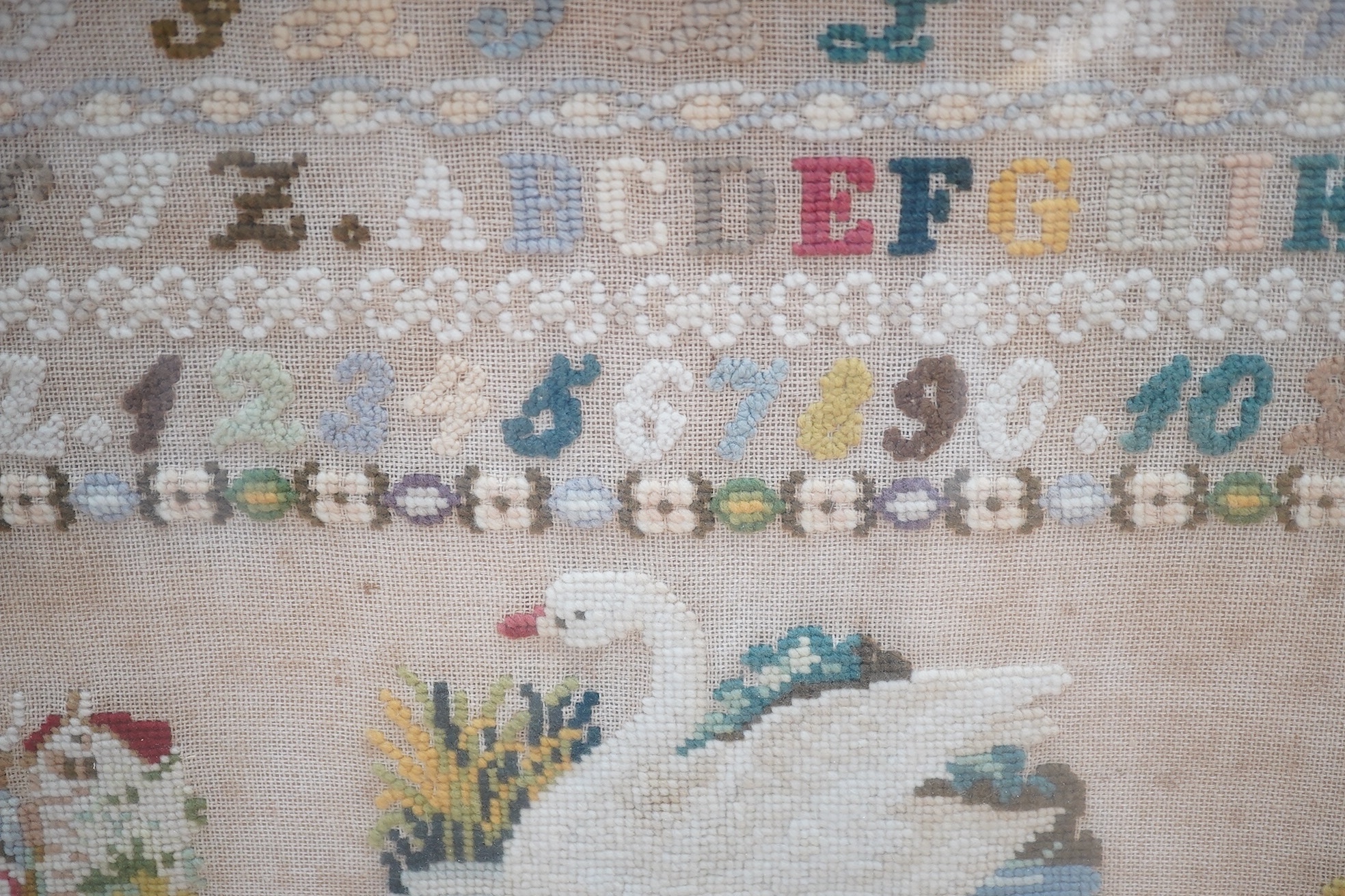 A framed sampler, initialled and dated 1874, worked with nine detailed pictorial motifs below a band of alphabet letters and numerals, each pictorial motif a different scene, a soldier and his wife, a white swan, a sailo